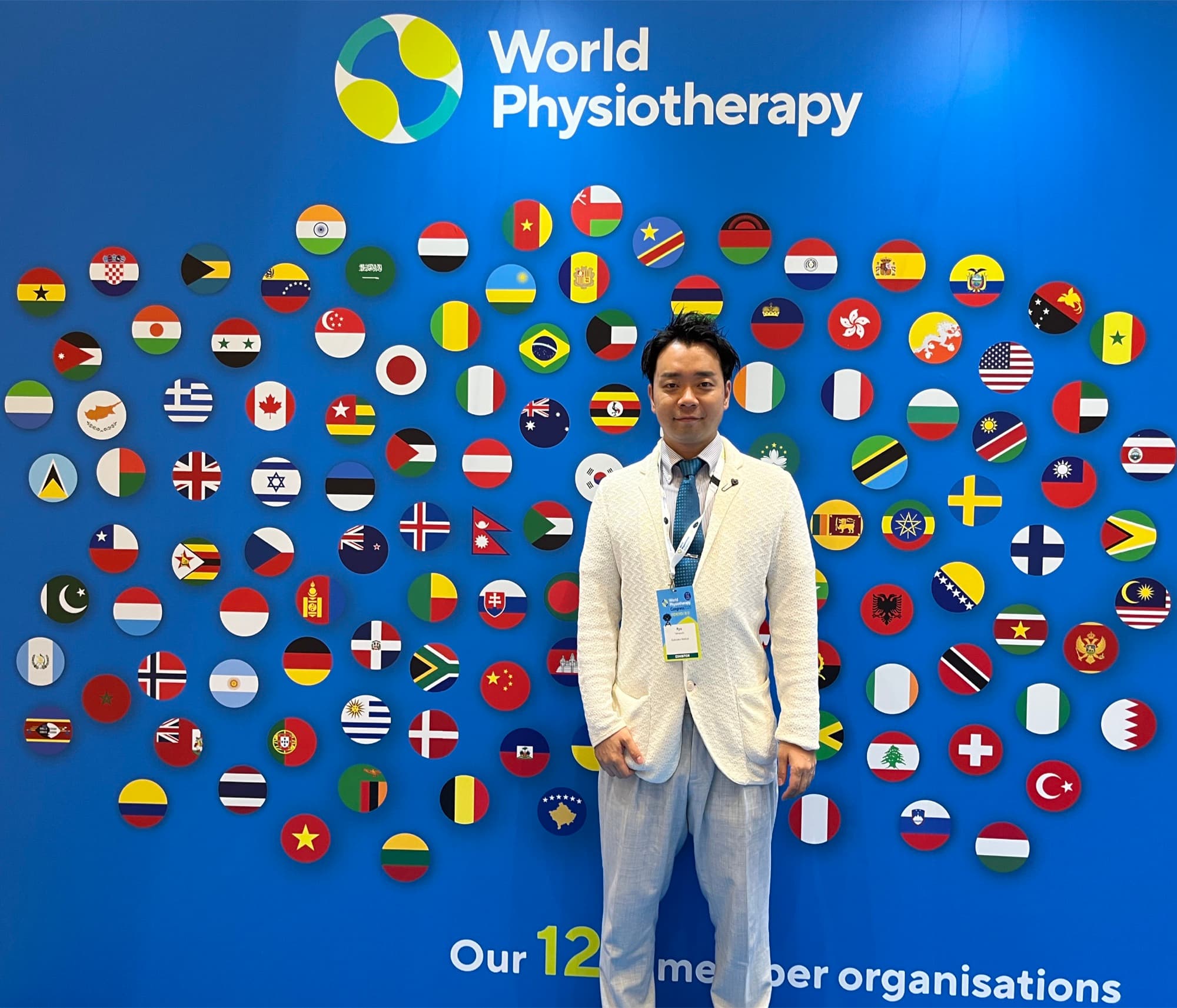 World Physiotherapyのロゴと世界の国旗が並ぶ会場フォトウォールの前で(出展者パス着用)
